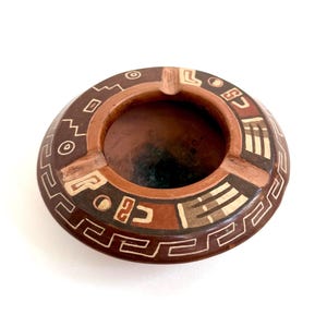 May include: A round, brown clay ashtray featuring a deep central basin and three notches on the rim. The exterior surface is decorated with cream-colored geometric patterns, including lines, circles, and angular shapes, set against a dark brown background.