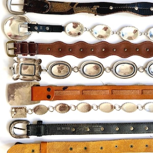 Op de afbeelding: Een collectie van acht riemen die horizontaal zijn gerangschikt. De riemen hebben diverse ontwerpen, waaronder donkerbruin leer met zilveren studs, zwart leer met ovale zilveren concho's, geschulpt bruin leer, zilveren schakelkettingen en effen leren banden.