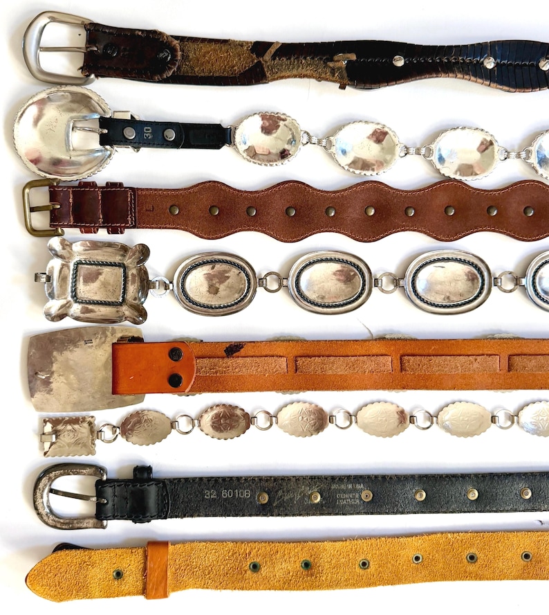 Op de afbeelding: Een collectie van acht riemen die horizontaal zijn gerangschikt. De riemen hebben diverse ontwerpen, waaronder donkerbruin leer met zilveren studs, zwart leer met ovale zilveren concho's, geschulpt bruin leer, zilveren schakelkettingen en effen leren banden.