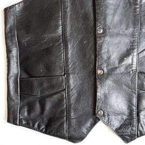Op de afbeelding: Een close-up van een zwart leren vest. Het vest heeft een klassiek ontwerp met een voorzak en drie knopen. Het leer heeft een licht versleten uiterlijk, wat bijdraagt aan de vintage uitstraling. Een klassiek kledingstuk.