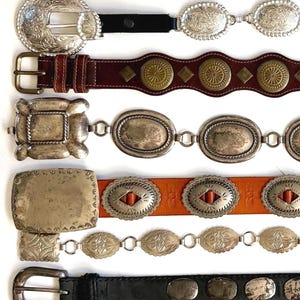 Op de afbeelding: Een collectie van acht riemen die horizontaal zijn gerangschikt. De riemen hebben diverse ontwerpen, waaronder leren banden met zilverkleurige gespen, metalen concho-accenten, messingkleurige ronde studs en rechthoekige metalen platen met gravures.
