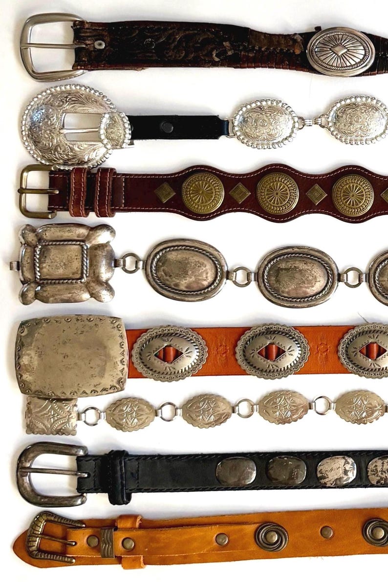 Op de afbeelding: Een collectie van acht riemen die horizontaal zijn gerangschikt. De riemen hebben diverse ontwerpen, waaronder leren banden met zilverkleurige gespen, metalen concho-accenten, messingkleurige ronde studs en rechthoekige metalen platen met gravures.
