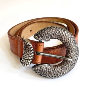Puede incluir: Un cinturón de cuero color canela con una hebilla circular grande de metal plateado con un patrón texturizado de escamas. La correa del cinturón es de cuero liso con costuras visibles en los bordes y una trabilla de metal plateado a juego.