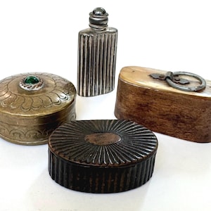 May include: A collection of antique boxes and a small bottle. The boxes include a round, brass-coloured box with a green gem, a dark brown box with a sunburst design, and a wooden box with a metal ring. The bottle is silver-coloured with vertical ridges.