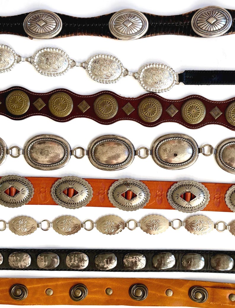 Op de afbeelding: Acht horizontaal gestapelde riemen. De riemen hebben diverse ontwerpen, waaronder zilverkleurige ovale concho's, messingkleurige ronde studs en gladde metalen platen op leren banden in tinten zwart, bruin en tan.