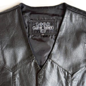 Op de afbeelding: Zwarte leren vest met een V-hals ontwerp. Het vest heeft een knoopsluiting en een label met de tekst "Leather Gallery" met maat "42". Het vest is gemaakt van een glanzend, getextureerd leermateriaal.