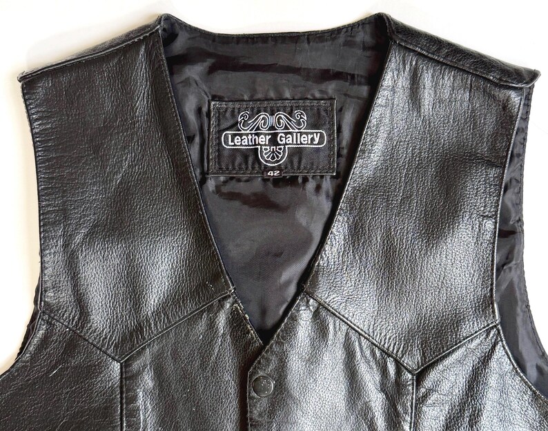 Op de afbeelding: Zwarte leren vest met een V-hals ontwerp. Het vest heeft een knoopsluiting en een label met de tekst "Leather Gallery" met maat "42". Het vest is gemaakt van een glanzend, getextureerd leermateriaal.