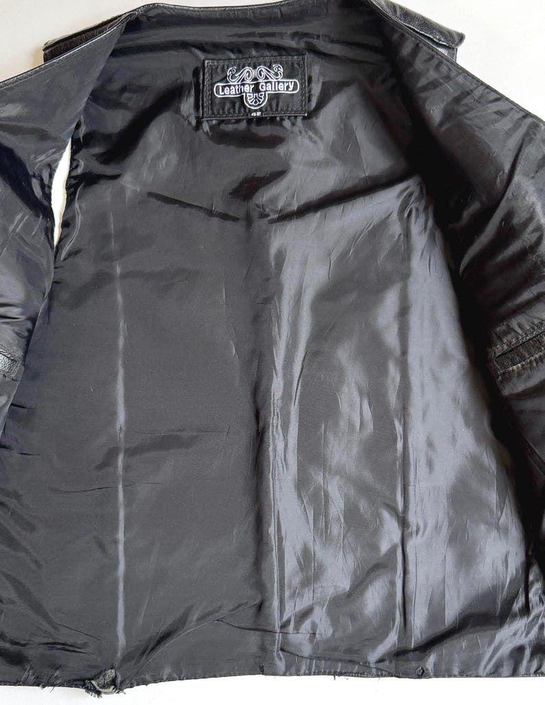 Op de afbeelding: Zwarte leren vest met een zwarte satijnen voering. De binnenkant heeft een label met de tekst "Leather Gallery". Het vest is open en toont de binnenvoering en stikdetails. Een klassiek bovenkledingstuk.