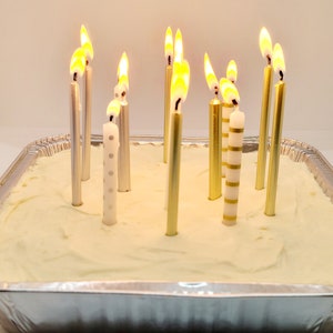 May include: A white frosted cake with 10 lit candles. The candles are silver and gold with white stripes. The cake is in a silver pan.