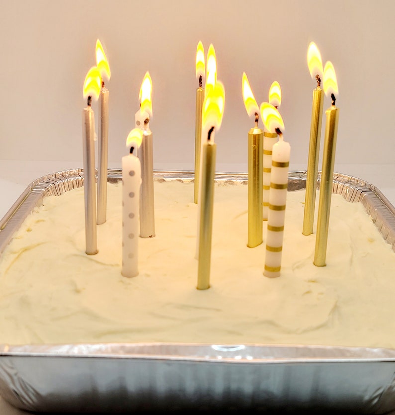 May include: A white frosted cake with 10 lit candles. The candles are silver and gold with white stripes. The cake is in a silver pan.