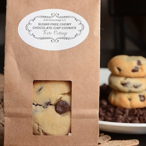 May include: A brown paper bag with a white label that reads "Sugar Free Chewy Chocolate Chip Cookies Keto Cottage" and a window showing a chocolate chip cookie.