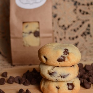May include: A stack of three chocolate chip cookies on a wooden surface. The cookies are light brown and have chocolate chips throughout. There are also chocolate chips scattered around the cookies.