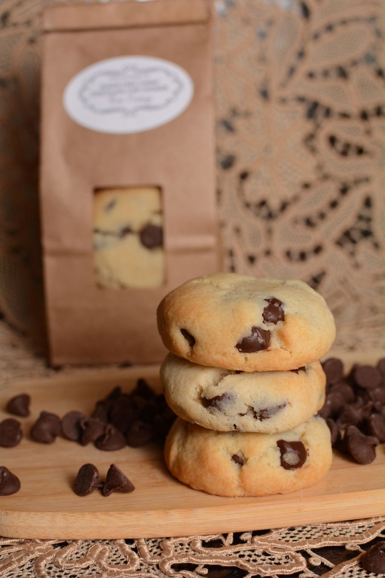 May include: A stack of three chocolate chip cookies on a wooden surface. The cookies are light brown and have chocolate chips throughout. There are also chocolate chips scattered around the cookies.