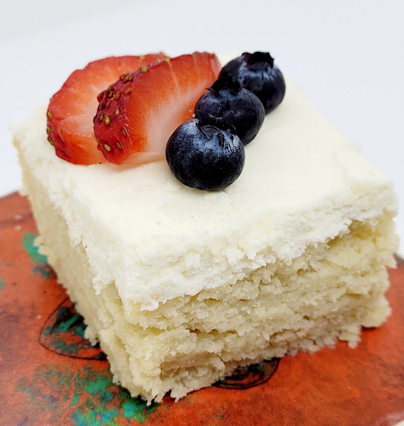 May include: A square slice of cake topped with white frosting and decorated with fresh strawberries and blueberries.