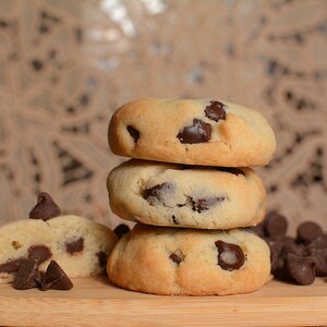 May include: A stack of three chocolate chip cookies on a wooden cutting board, with a broken cookie in the foreground and chocolate chips scattered around.