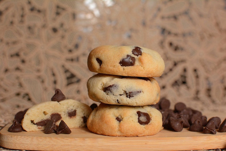 May include: A stack of three chocolate chip cookies on a wooden cutting board, with a broken cookie in the foreground and chocolate chips scattered around.