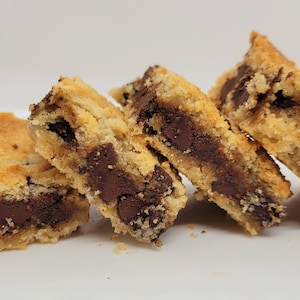 May include: Four chocolate chip cookie bars stacked on a white plate. The bars are made with a golden brown cookie dough and filled with dark chocolate chips.