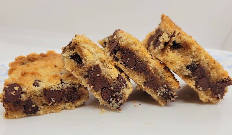May include: Four chocolate chip cookie bars stacked on a white plate. The bars are made with a golden brown cookie dough and filled with dark chocolate chips.