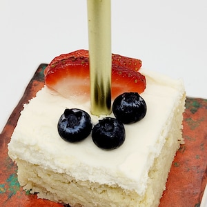 May include: A single-layer square cake with white frosting, topped with fresh strawberries and blueberries. A gold candle is inserted into the frosting.