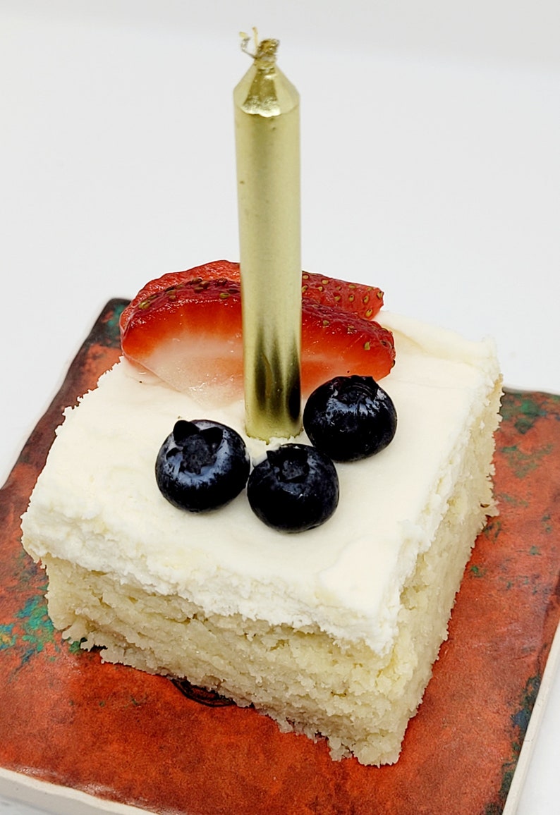May include: A single-layer square cake with white frosting, topped with fresh strawberries and blueberries. A gold candle is inserted into the frosting.