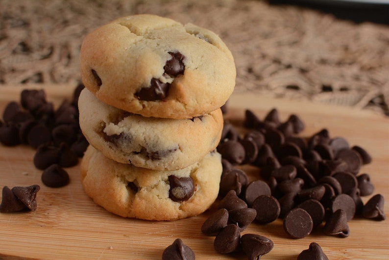 May include: Three chocolate chip cookies stacked on top of each other on a wooden surface. The cookies are light brown and have chocolate chips throughout. There are chocolate chips scattered around the cookies.