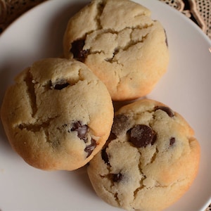 May include: Three golden brown chocolate chip cookies on a white plate. The cookies have a slightly crispy exterior and a soft, chewy interior.