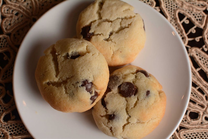 May include: Three golden brown chocolate chip cookies on a white plate. The cookies have a slightly crispy exterior and a soft, chewy interior.
