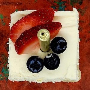 May include: A white square cake topped with strawberries and blueberries. A gold candle is in the center of the cake.