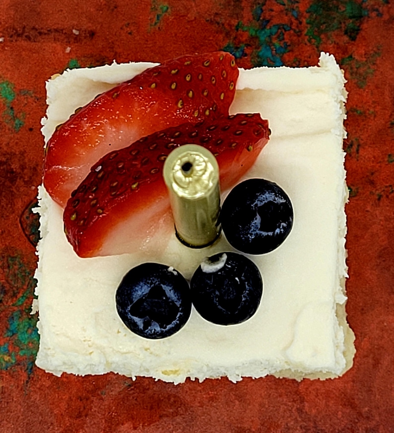 May include: A white square cake topped with strawberries and blueberries. A gold candle is in the center of the cake.
