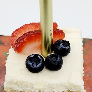 May include: A single-layer square cake with white frosting, topped with fresh strawberries, blueberries, and a gold candle.