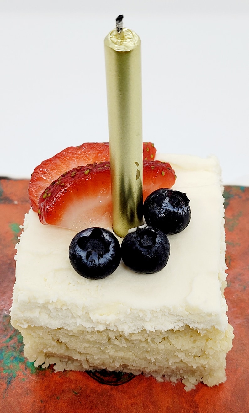 May include: A single-layer square cake with white frosting, topped with fresh strawberries, blueberries, and a gold candle.