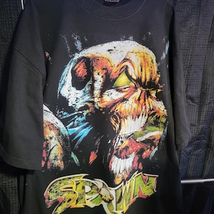 May include: Black t-shirt featuring a vibrant graphic of a character with a skull-like face, sharp teeth, and a fiery appearance. The word "SPAWN" is displayed in a stylized font. The shirt is hanging on a wooden hanger.