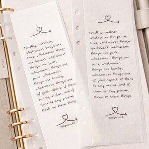 May include: Two clear bookmarks with handwritten text from Philippians 4:8, featuring a heart and arrow design. The bookmarks are inside a beige planner with gold accents.