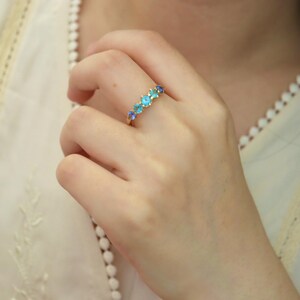 May include: A gold ring with a row of blue and green gemstones. The ring is on a person's finger.