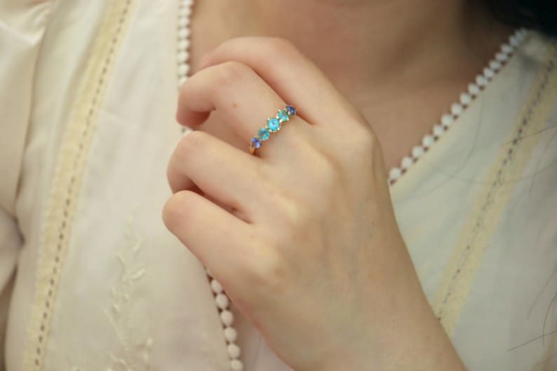 May include: A gold ring with a row of blue and green gemstones. The ring is on a person's finger.