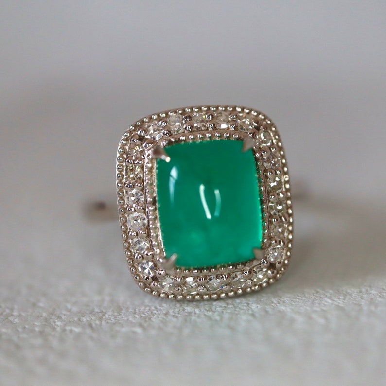 May include: A green gemstone ring with a square-shaped setting surrounded by a halo of small diamonds.