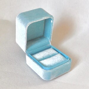 May include: A light blue velvet ring box, open to reveal two ring slots. The box is square with rounded edges and a contrasting light blue interior. The velvet material gives a soft, luxurious appearance.