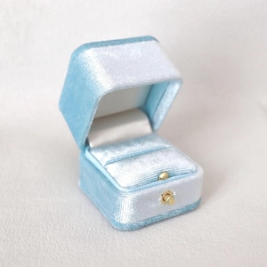 May include: Open, light blue velvet ring box with a white interior. The box is rectangular with a hinged lid and a gold-colored clasp. The interior has a slot to hold a ring.