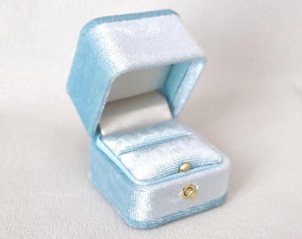 Könnte beinhalten: Geöffnete, hellblaue Samt-Ringbox mit weißem Innenraum. Die rechteckige Box hat einen Klappdeckel und einen goldfarbenen Verschluss. Im Inneren befindet sich ein Schlitz zur Aufnahme eines Rings.