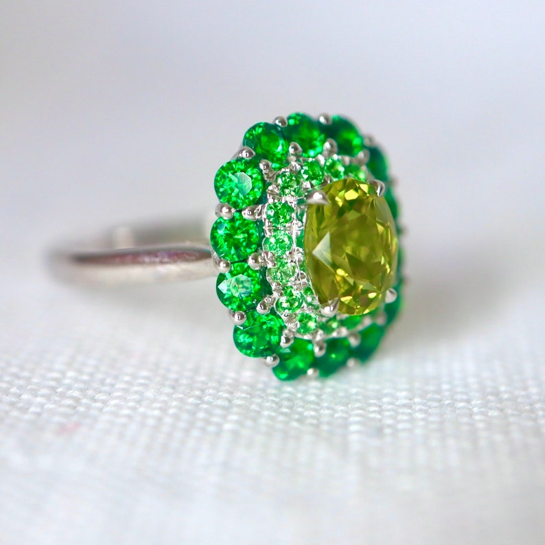 May include: A silver ring with a large, oval-shaped green gemstone surrounded by a double halo of smaller green gemstones.