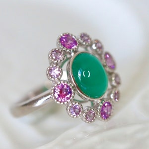 May include: A silver ring with a green gemstone surrounded by a halo of pink gemstones.