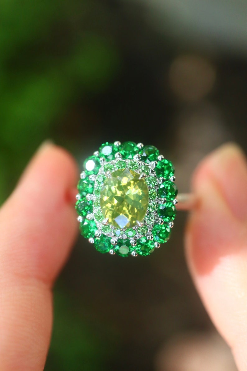 May include: A ring with a large, oval-shaped, light green gemstone surrounded by a double halo of smaller green gemstones.