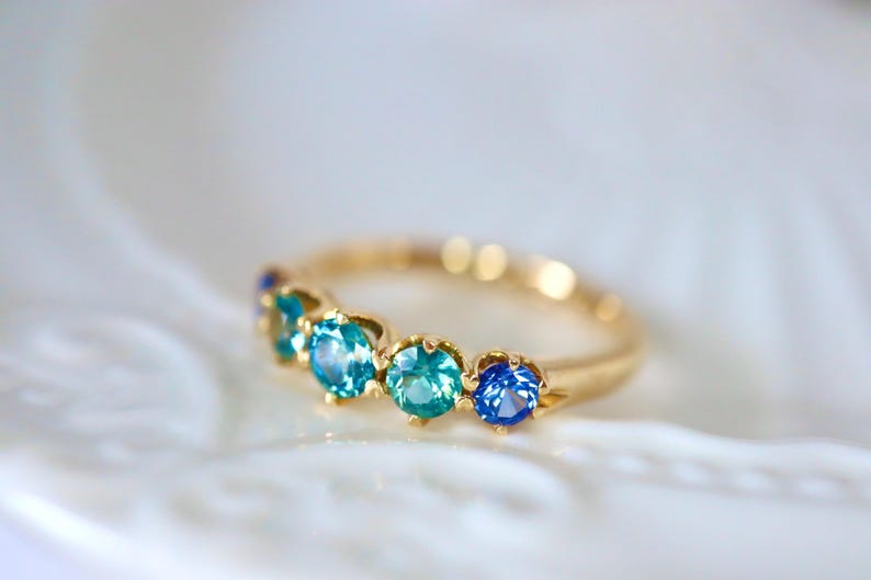 May include: A gold band ring with five blue gemstones. The gemstones are set in a curved line and are different shades of blue.