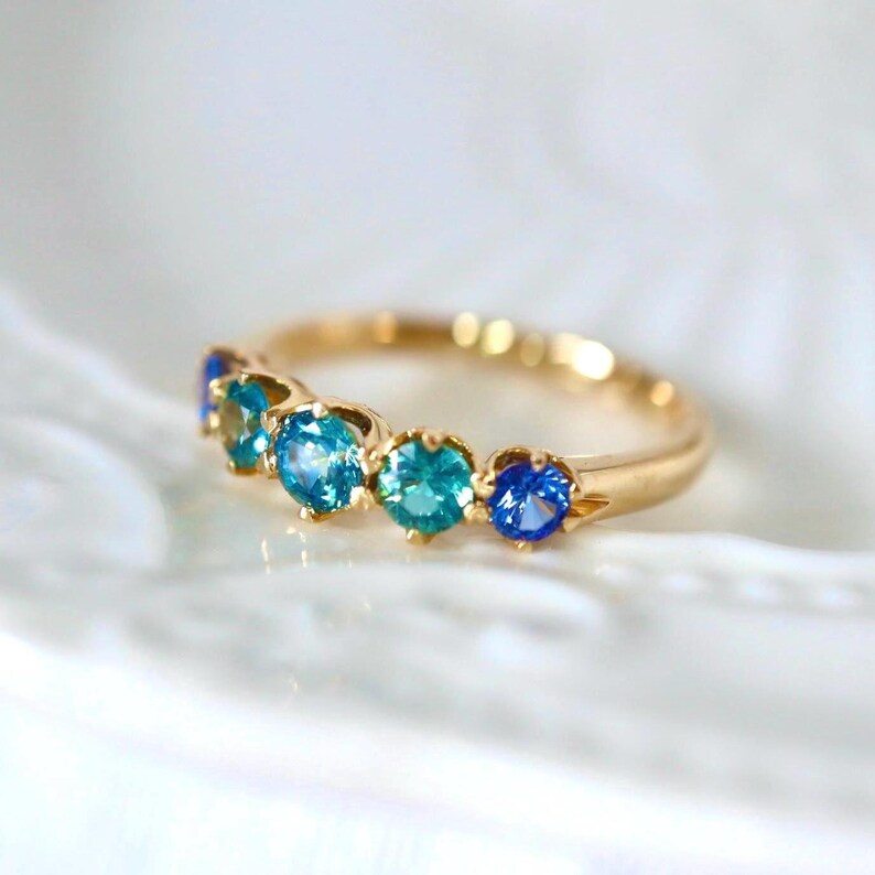 May include: A gold band ring with six gemstones. The gemstones are a gradient of blue and green colors.