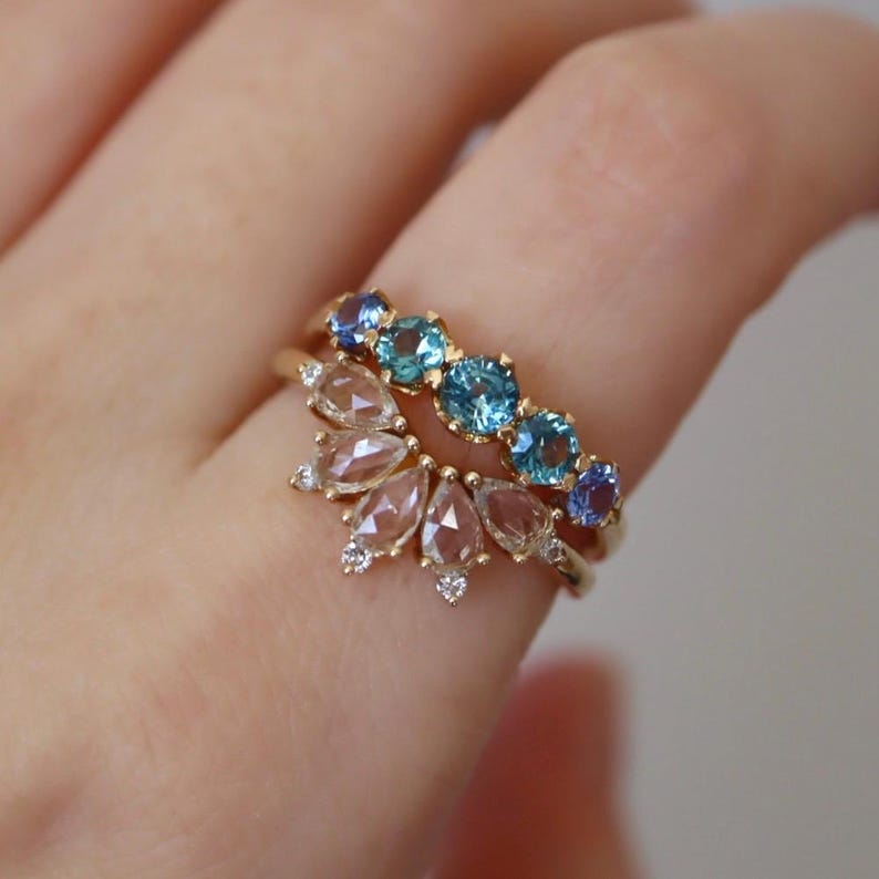 May include: Two gold rings with gemstones. The top ring features a row of blue sapphires. The bottom ring features a cluster of pear-shaped diamonds.