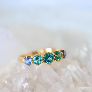May include: A gold band ring with a row of blue and green gemstones. The ring is set on a white background.