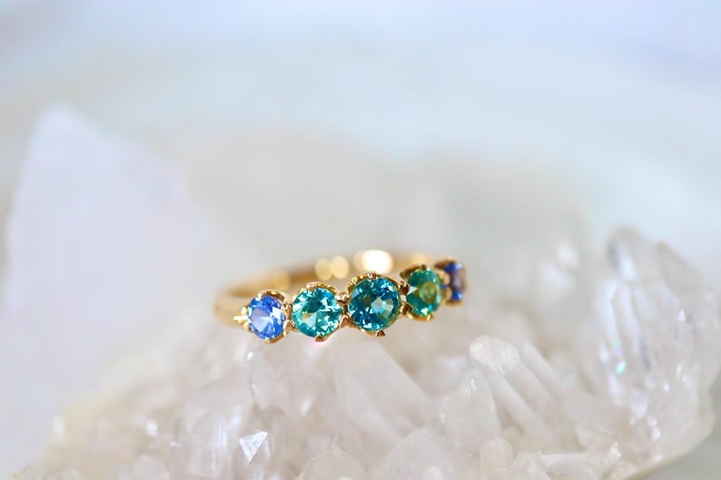 May include: A gold band ring with a row of blue and green gemstones. The ring is set on a white background.