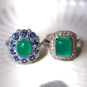 May include: Two silver rings with green gemstones and blue sapphires. The ring on the left has a larger oval-shaped green gemstone surrounded by smaller blue sapphires and diamonds. The ring on the right has a square-shaped green gemstone surrounded by diamonds.