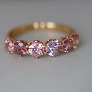May include: A gold ring featuring a series of pink gemstones. The gemstones are round-cut and set closely together, creating a continuous band of color. The ring's band is a warm gold tone, and the gemstones vary in shades of pink.