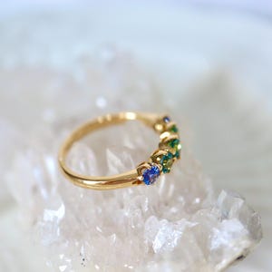 May include: A gold band ring with a row of blue and green gemstones. The ring is set on a white crystal surface.